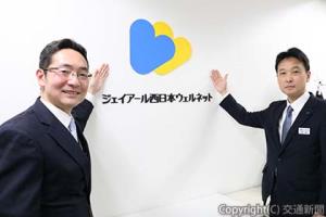 フロア入口横の社名ロゴの前で記念撮影する吉田社長（左）と荒武理事（ジェイアール西日本ウェルネット提供）